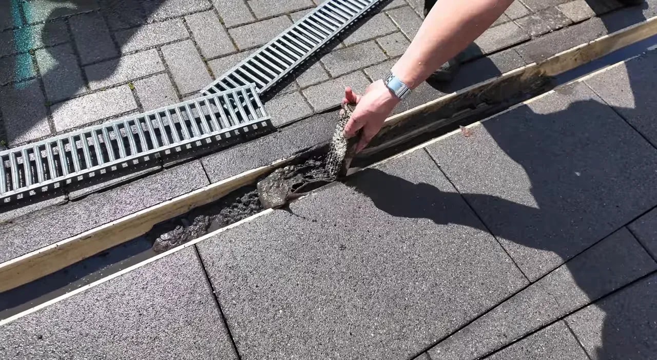 Hand entfernt Schmutz und Ablagerungen aus einer geöffneten Abflussrinne zwischen Steinplatten und Pflaster