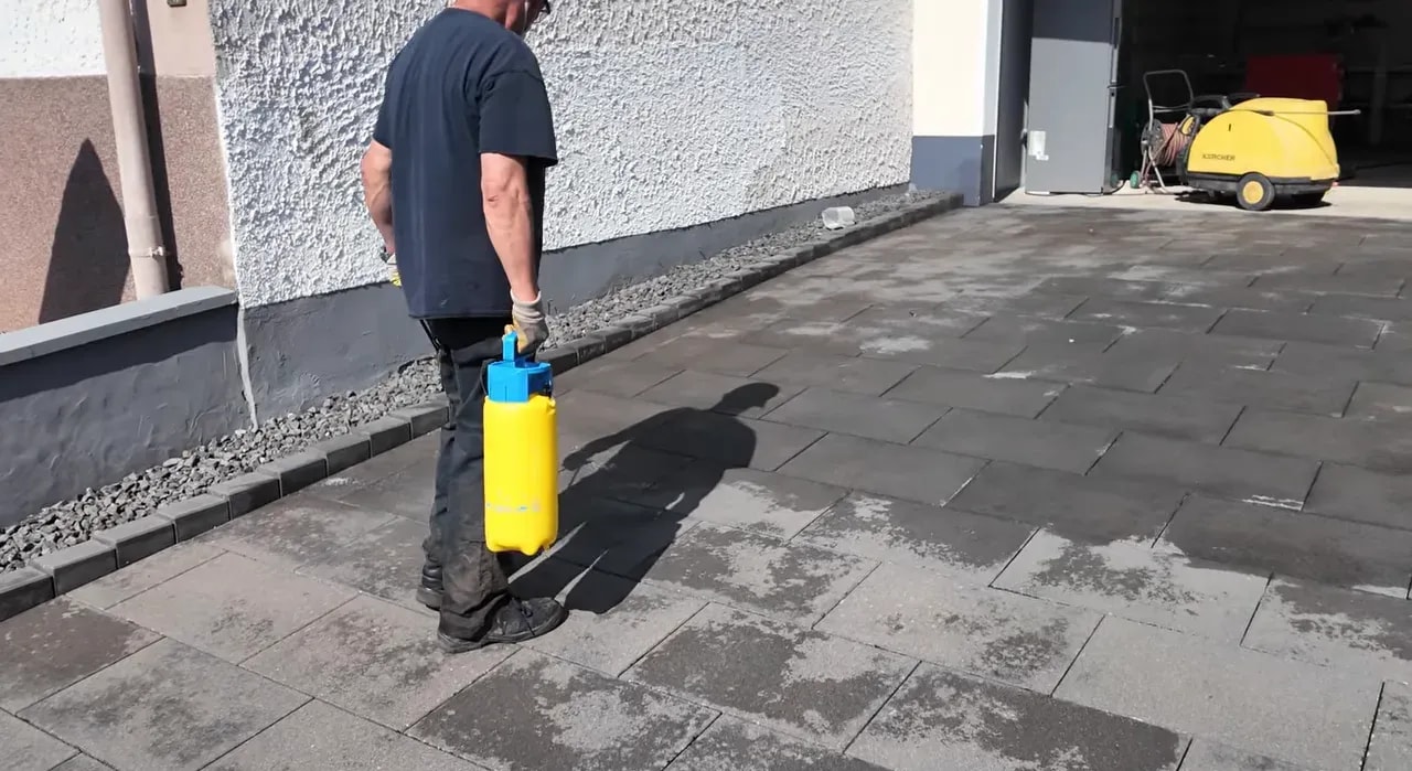 Person läuft mit gelber Druckspritze über eine gereinigte Einfahrt und trägt Reiniger auf